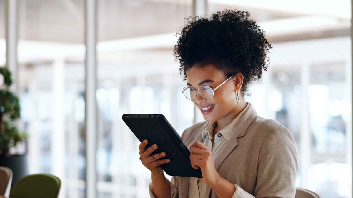 Junge Frau mit Brille steht auf der Arbeit und hält ein Tablet in der Hand.