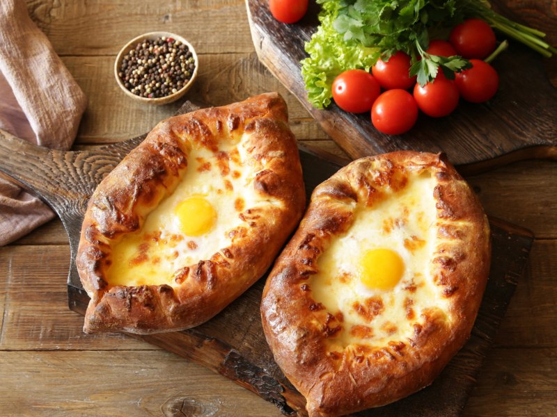 Zwei Khachapuri auf einem Holzbrett auf Esstisch, Teigboote mit Käse und Ei-Füllung.