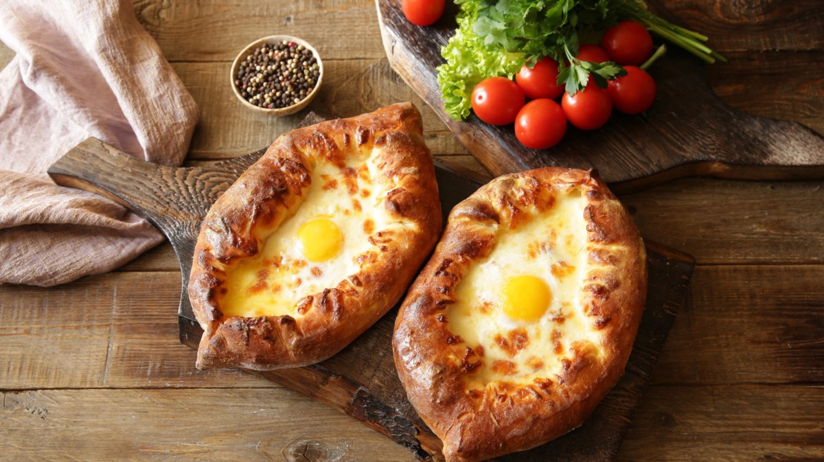 Zwei Khachapuri auf einem Holzbrett auf Esstisch, Teigboote mit Käse und Ei-Füllung.