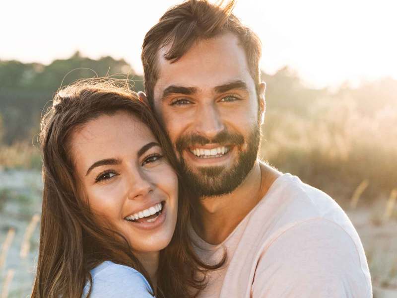 Mann und Frau mit braunen Haaren stehen eng beieinander am Strand und lachen.