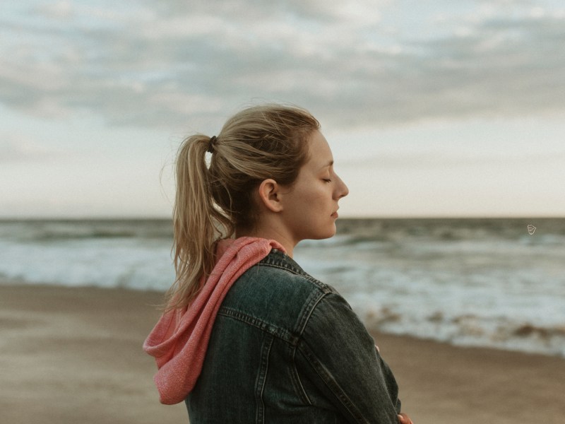 Frau am Strand mit Wellen im Hintergrund, die nachdenklich nach vorne blickt.