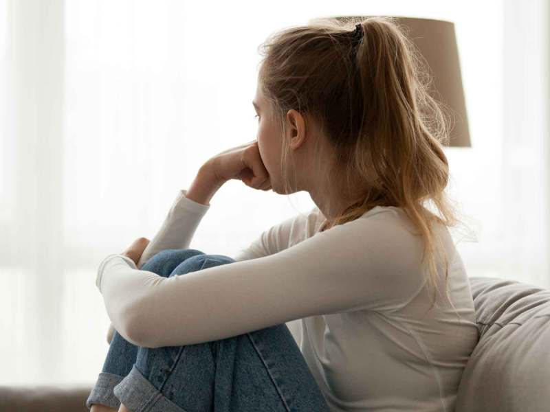 Junge Frau mit weißem Shirt sitzt nachdenklich auf der Couch und blickt aus dem Fenster.