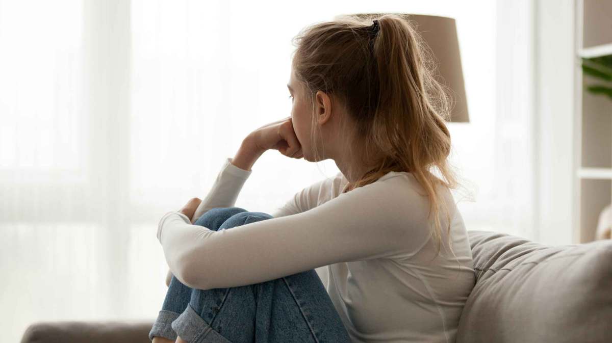 Junge Frau mit weißem Shirt sitzt nachdenklich auf der Couch und blickt aus dem Fenster.