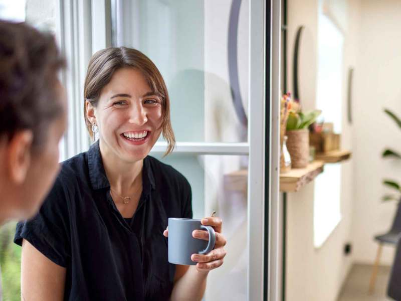 Frau sitzt auf einer Fensterbank mit einer Kollegin und lacht.