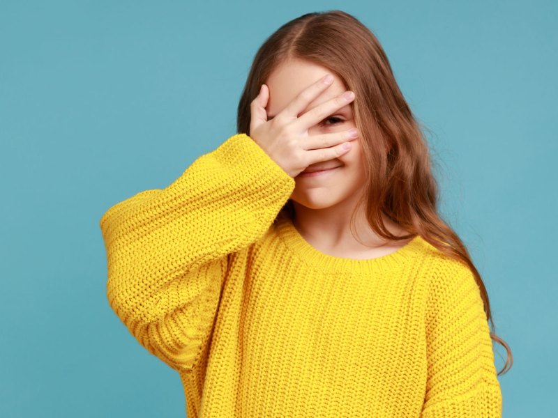 Mädchen, ca 10 Jahre alt, steht mit einem gelben Pullover vor einer türkisen Wand und versteckt ihr Gesicht hinter der rechten Hand.