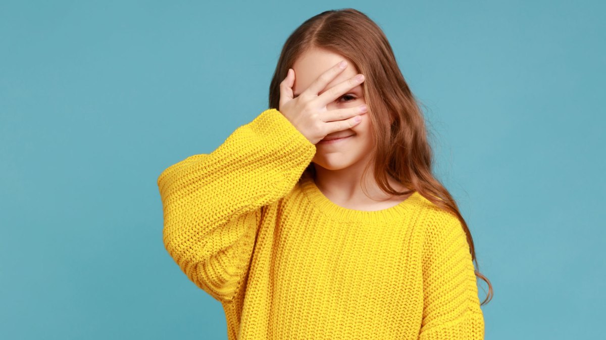 Mädchen, ca 10 Jahre alt, steht mit einem gelben Pullover vor einer türkisen Wand und versteckt ihr Gesicht hinter der rechten Hand.