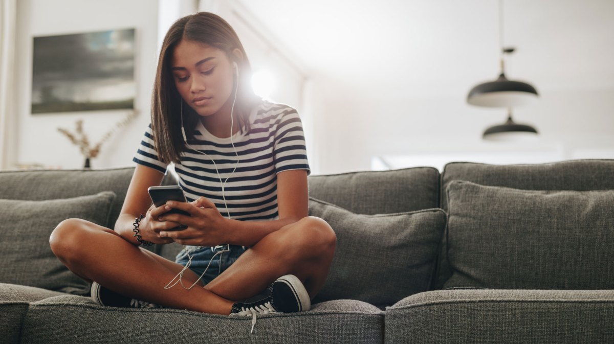 Mädchen im Teenageralter sitzt auf dem Sofa und hört etwas über das Handy mit Kopfhörern.