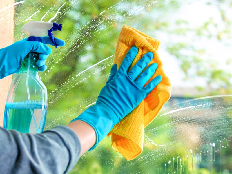 Person putzt Fenster, hält gelbes Tuch in einer Hand und Sprühflasche mit Reiniger in anderer.