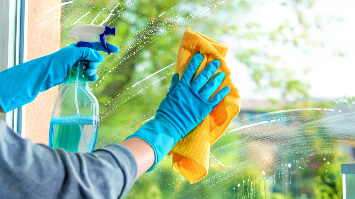 Person putzt Fenster, hält gelbes Tuch in einer Hand und Sprühflasche mit Reiniger in anderer.