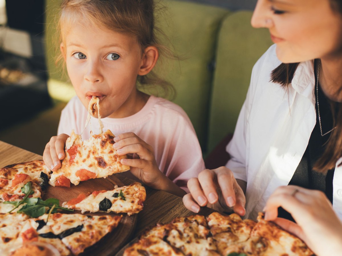 Mutter und Tochter sitzen in einem Cafe und jede hat eine Pizza vor sich auf dem Teller. Das Mädchen hat gerade von einem Stück abgebissen.