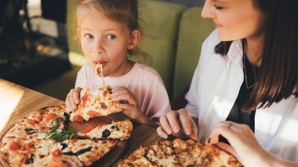 Mutter und Tochter sitzen in einem Cafe und jede hat eine Pizza vor sich auf dem Teller. Das Mädchen hat gerade von einem Stück abgebissen.