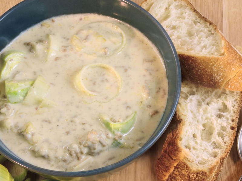 Käse-Hack-Lauch-Suppe in einer schwarzen Schüssel, daneben liegen Bageuttebrot und ein Löffel.