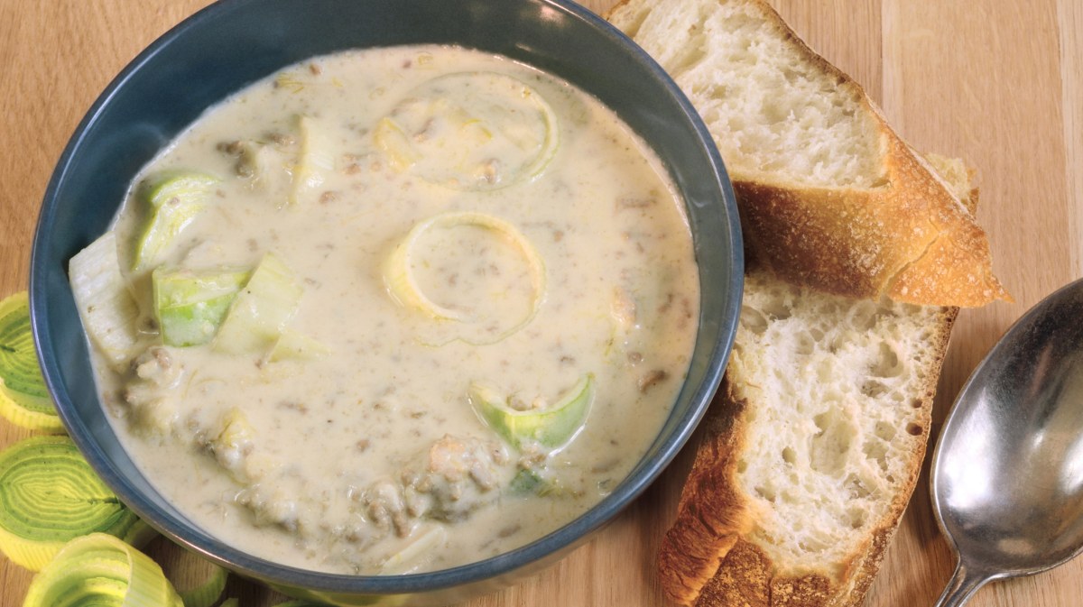 Käse-Hack-Lauch-Suppe in einer schwarzen Schüssel, daneben liegen Bageuttebrot und ein Löffel.