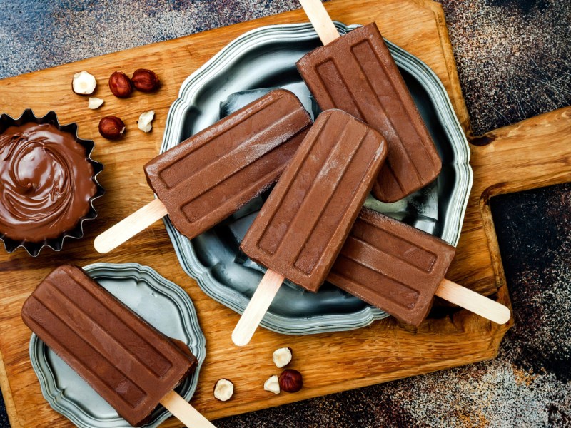 Schokoladen-Stieleis auf einem Holzbrett, daneben eine kleine Schüssel mit Schokocreme.