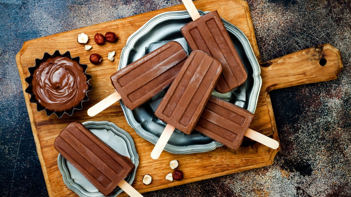 Schokoladen-Stieleis auf einem Holzbrett, daneben eine kleine Schüssel mit Schokocreme.