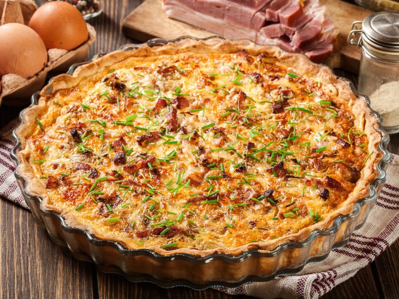 Quiche Lorraine in Form auf einem Holztisch, daneben liegen Eier und Speck.