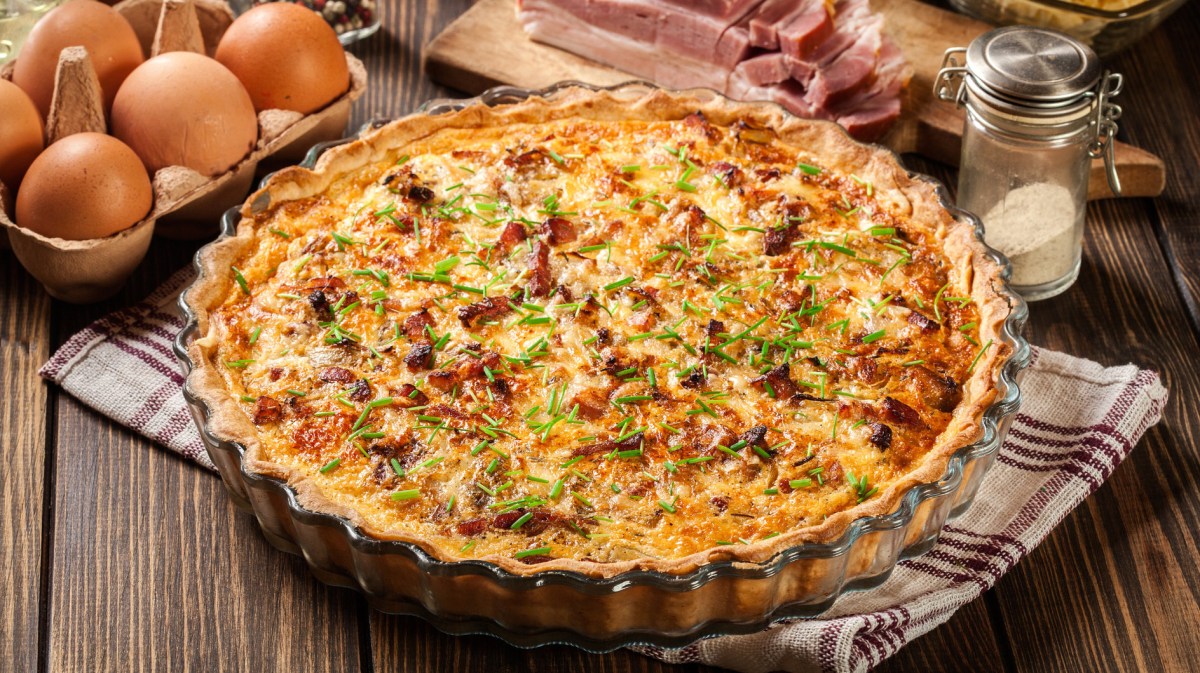 Quiche Lorraine in Form auf einem Holztisch, daneben liegen Eier und Speck.