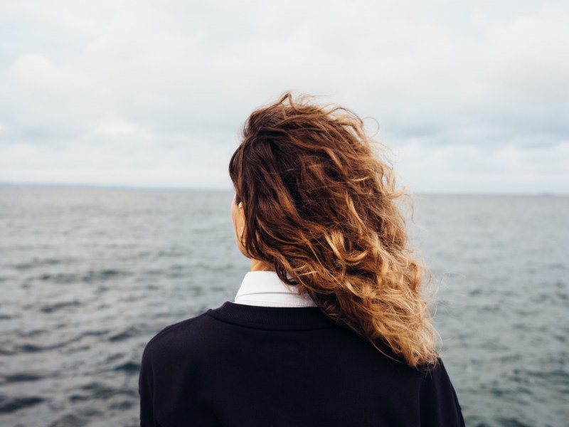 Frau mit Locken hat Rücken zur Kamera gewandt und schaut aufs Meer hinaus.