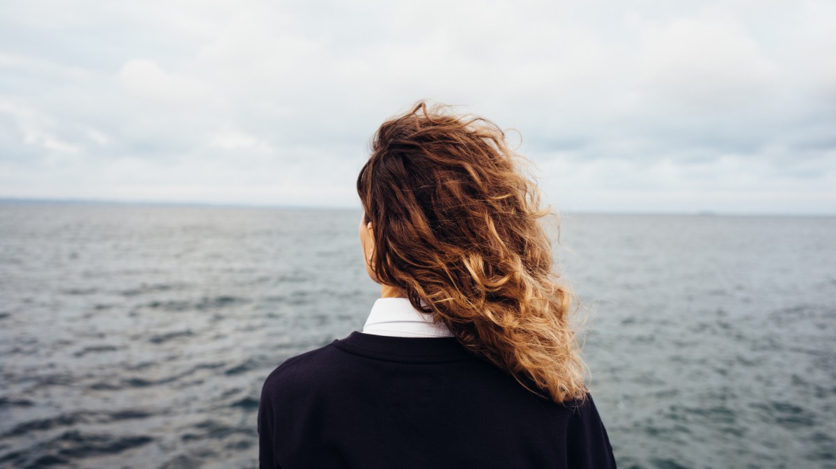 Frau mit Locken hat Rücken zur Kamera gewandt und schaut aufs Meer hinaus.