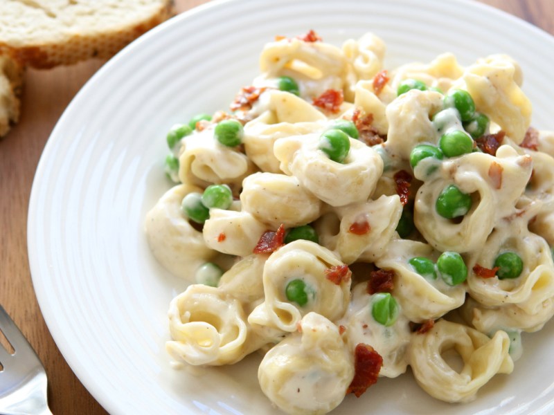 Tortellini mit heller Soße, Speck und Erbsen auf einem weißen Teller.