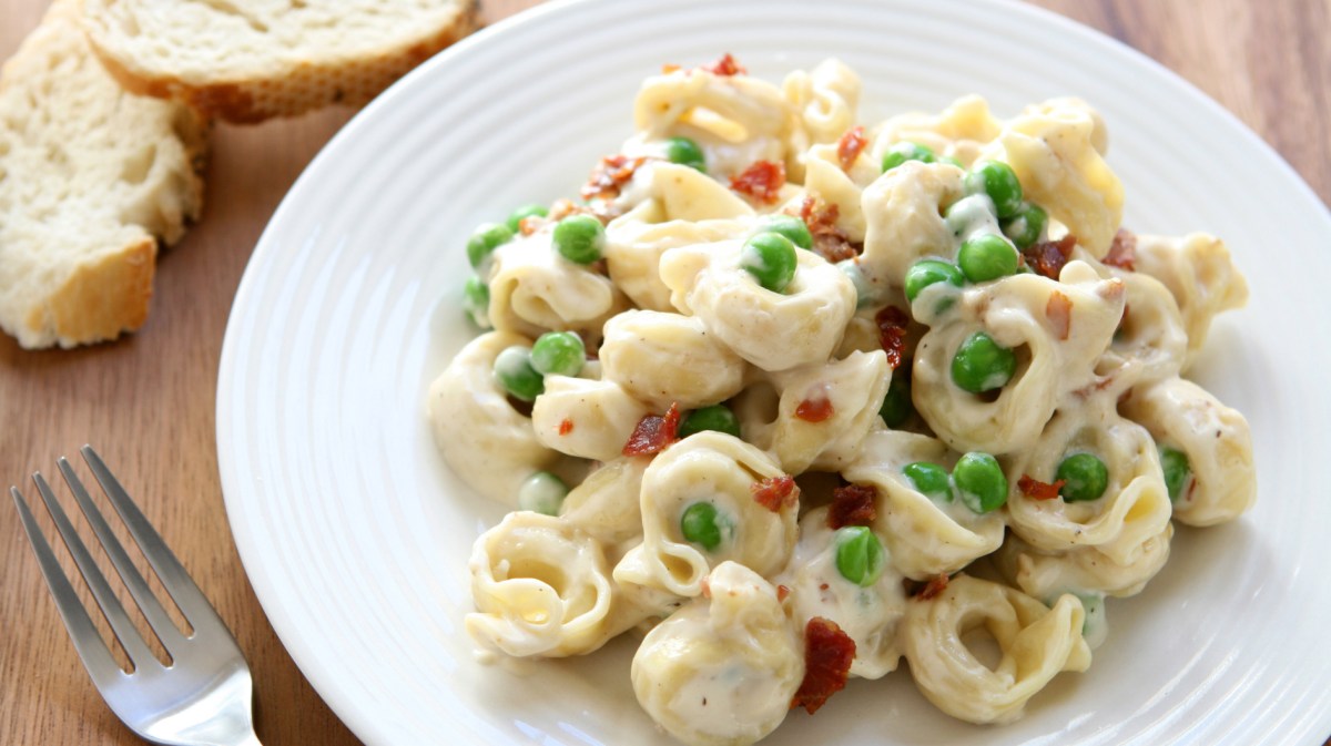 Tortellini mit heller Soße, Speck und Erbsen auf einem weißen Teller.