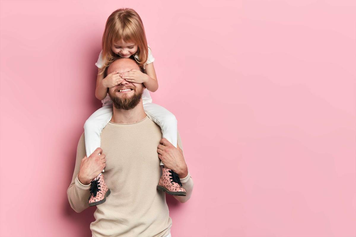 Vater trägt Tochter auf den Schultern vor einer rosa Wand. Sie hält ihm die Augen zu.