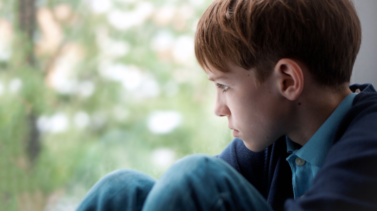 Nahaufnahme eines Jungen, der an einem Fenster sitzt und niedergeschlagen nach draußen sieht.