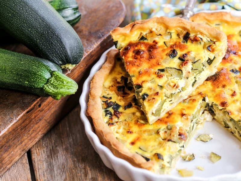 Quiche mit Zucchini und Feta in einer weißen Form, daneben liegen Zucchini.