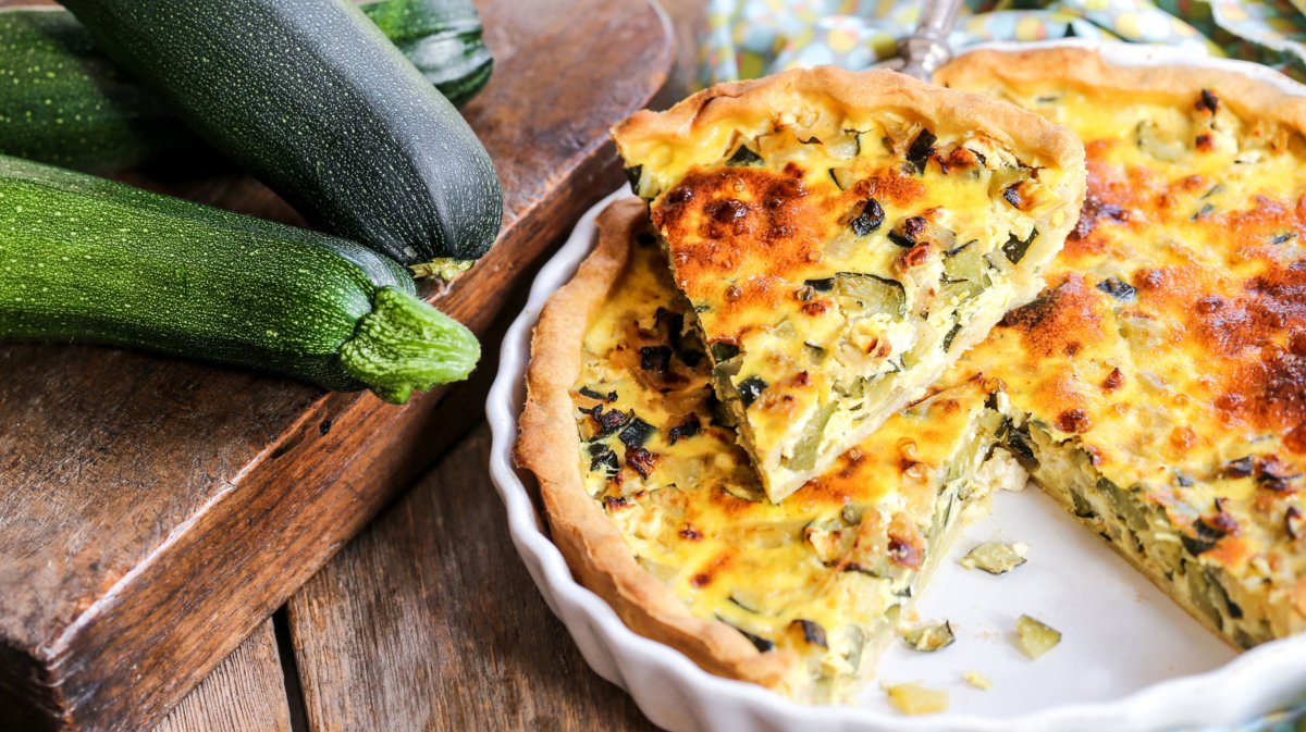 Quiche mit Zucchini und Feta in einer weißen Form, daneben liegen Zucchini.