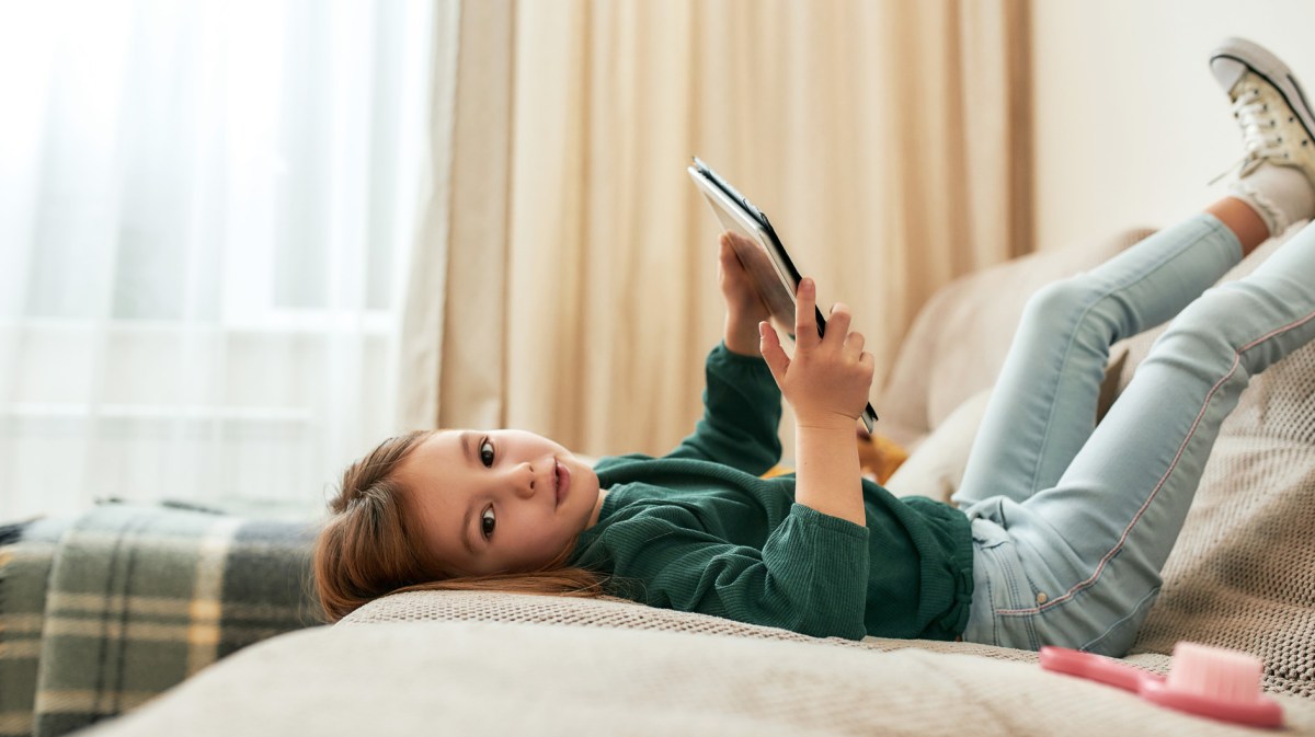 Mädchen, ca. 6 Jahre alt, liegt mit Tablet auf dem Sofa, die Beine in die Luft gestreckt.