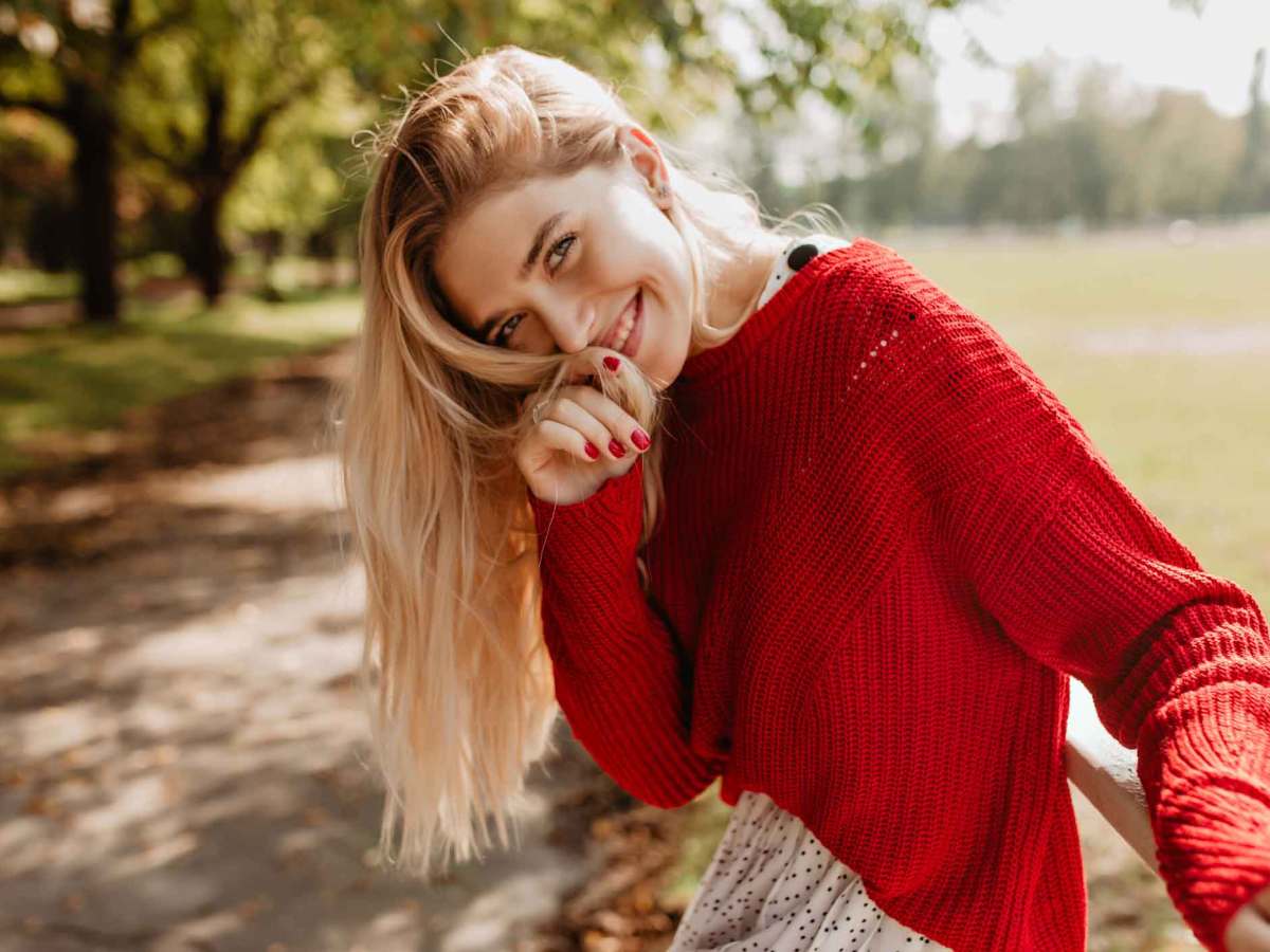 Junge Frau mit blonden Haaren und einem roten Pullover lächelt in die Kamera und sitzt draußen.