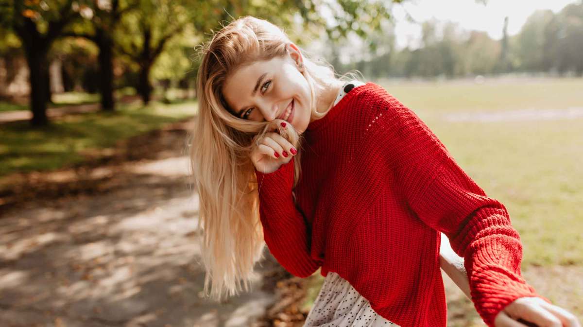 Junge Frau mit blonden Haaren und einem roten Pullover lächelt in die Kamera und sitzt draußen.