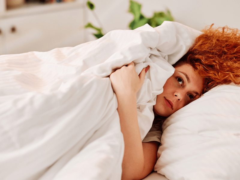 Frau mit roten Haaren im bett, die sich unter der Bettdecke versteckt.