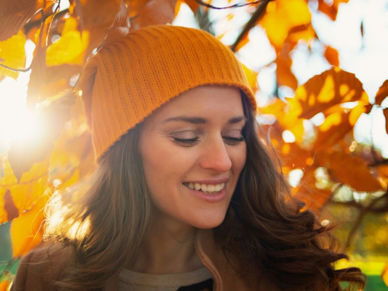 Frau mit Mütze bei Sonnenschein vor einem herbstlichen Laubbaum.