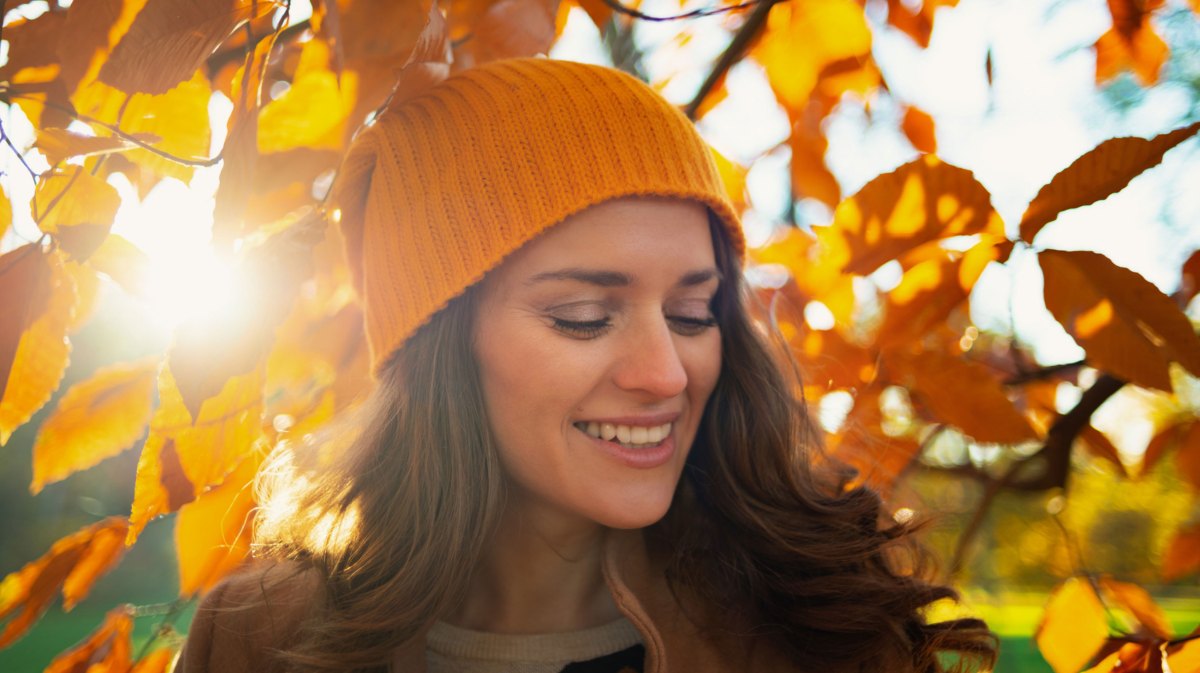 Frau mit Mütze bei Sonnenschein vor einem herbstlichen Laubbaum.