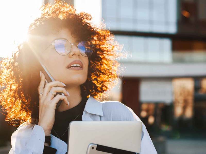 Junge Frau mit Brille und Locken telefoniert beruflich und hat ein Tablet im Arm.