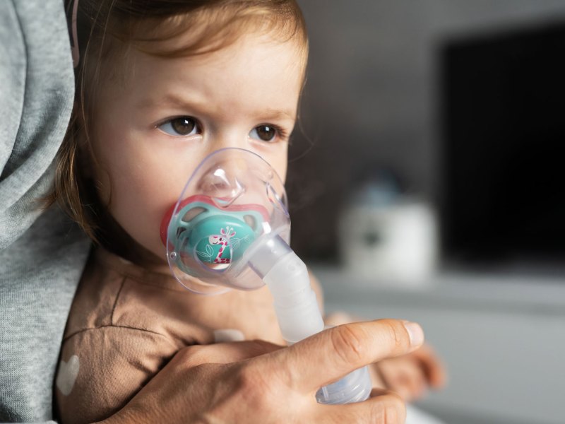 Kleines Mädchen, ca. 2 Jahre alt, sitzt auf dem Schoß eines Erwachsenen und atmet in eine Inhalator-Maske.