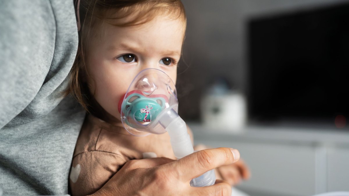 Kleines Mädchen, ca. 2 Jahre alt, sitzt auf dem Schoß eines Erwachsenen und atmet in eine Inhalator-Maske.