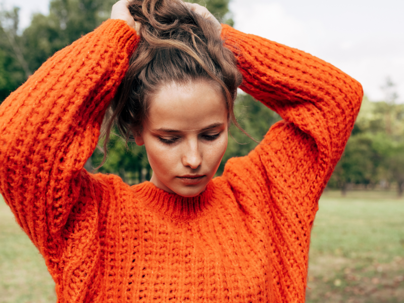 Frau mit rotem Pulli auf einem Rasen, die sich nachdenklich durch die Haare geht.