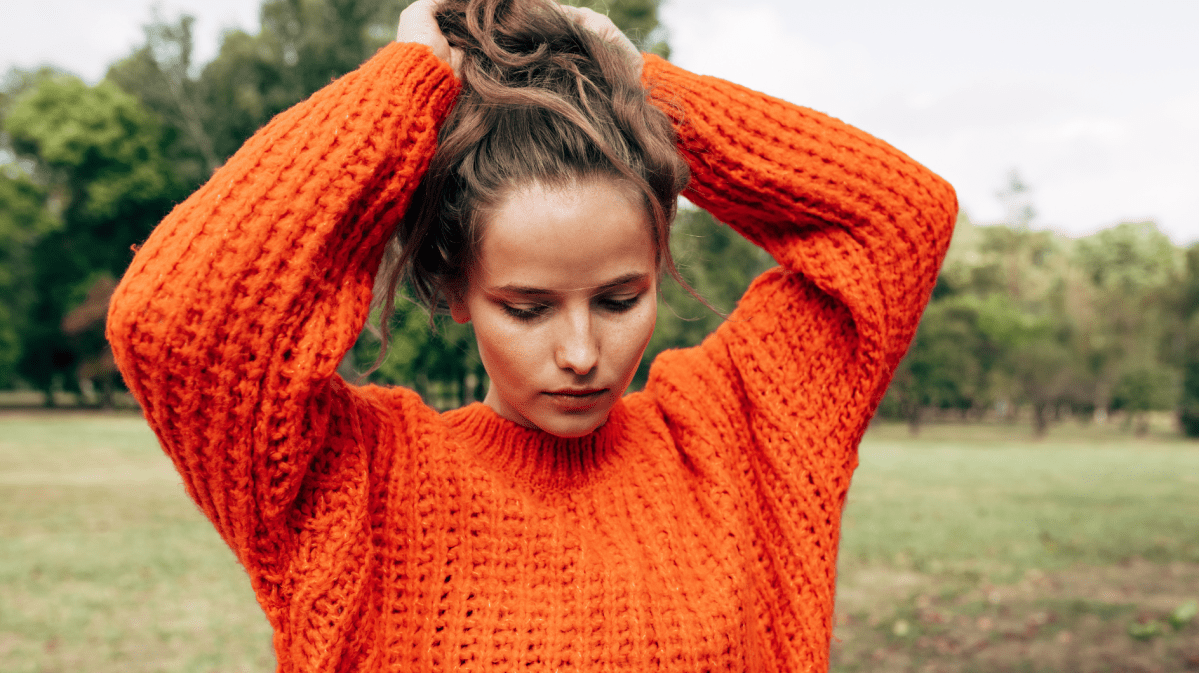 Frau mit rotem Pulli auf einem Rasen, die sich nachdenklich durch die Haare geht.