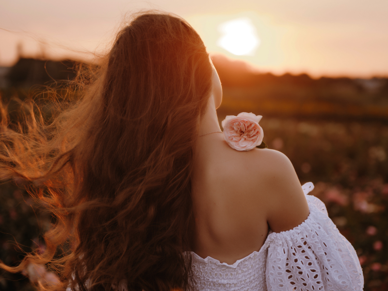 Frau auf einem Feld in einem weißen Kleid mit Blume in der Hand, die sich mit dem Rücken zur Kamera dreht.