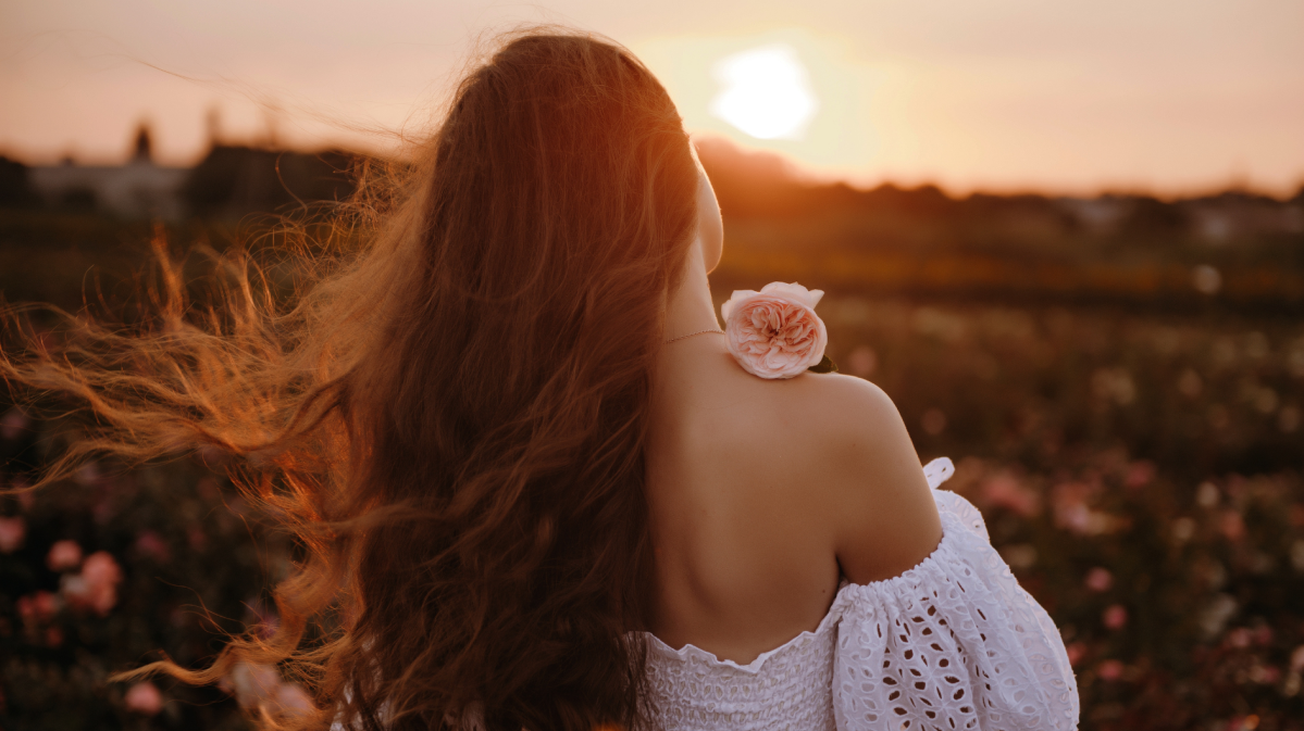 Frau auf einem Feld in einem weißen Kleid mit Blume in der Hand, die sich mit dem Rücken zur Kamera dreht.