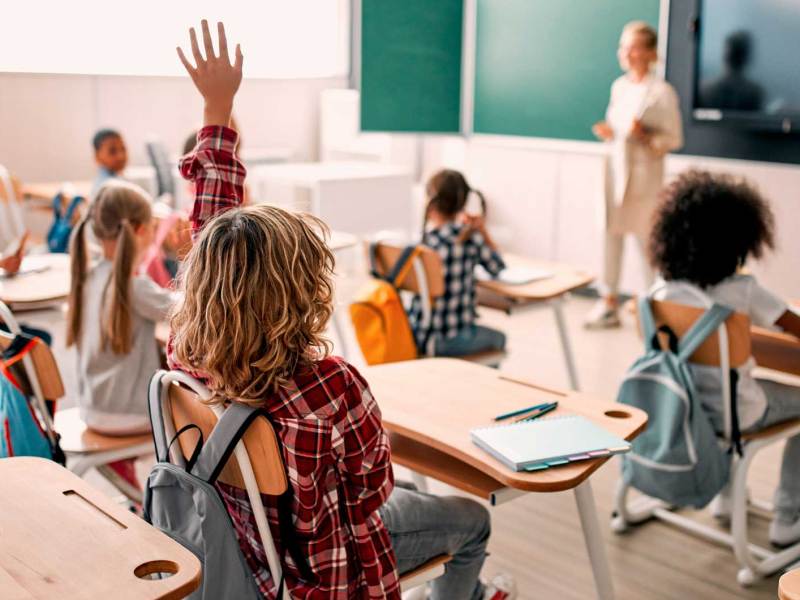 Blick in ein Klassenzimmer. Die Kinder heben zum Teil die Hände, die Lehrerin steht vorn vor der Tafel, mit dem Blick ins Klassenzimmer.