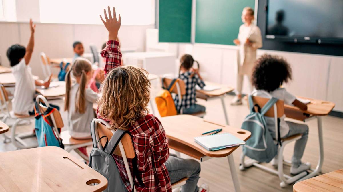 Blick in ein Klassenzimmer. Die Kinder heben zum Teil die Hände, die Lehrerin steht vorn vor der Tafel, mit dem Blick ins Klassenzimmer.