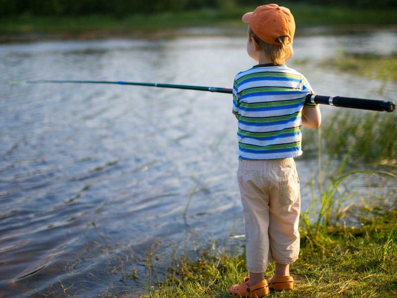 Junge, ca. 7 Jahre, steht mit einer Angel in der Hand an einem See und wartet darauf, dass ein Fisch anbeißt.