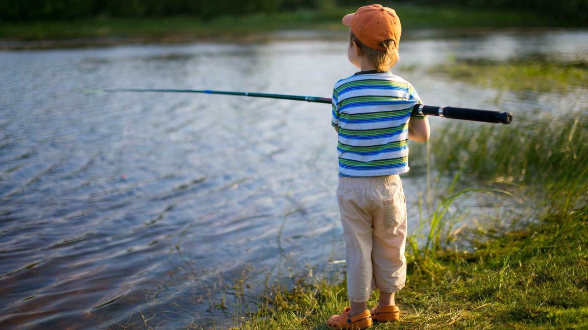 Junge, ca. 7 Jahre, steht mit einer Angel in der Hand an einem See und wartet darauf, dass ein Fisch anbeißt.