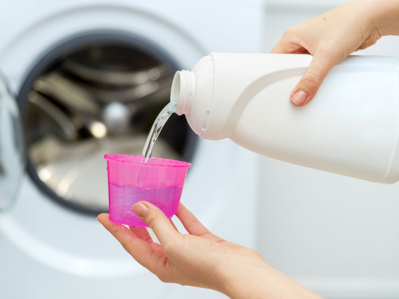 Hände die Waschmaschinenreiniger aus weißer Flasche in rosa Becher füllen, dahinter Waschmaschine