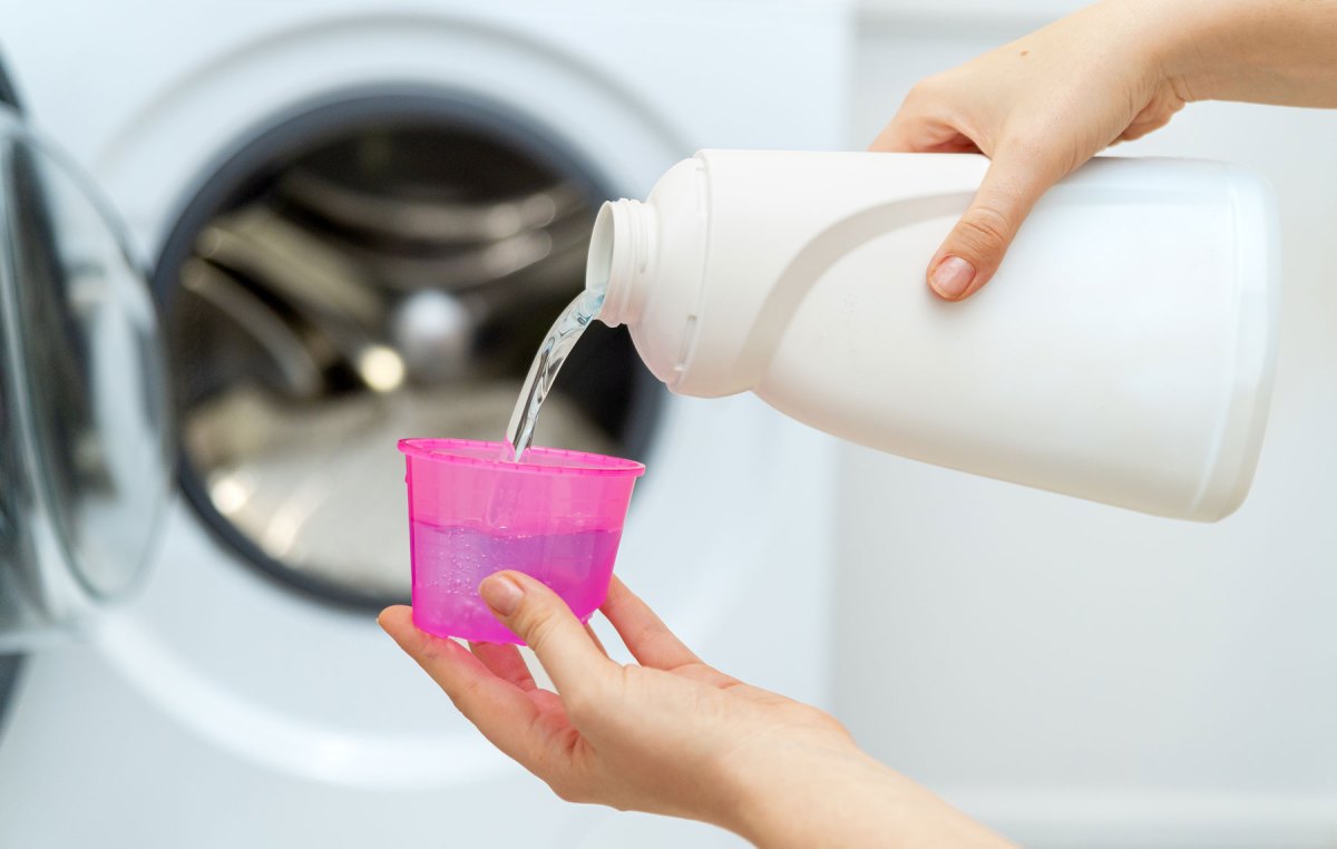 Hände die Waschmaschinenreiniger aus weißer Flasche in rosa Becher füllen, dahinter Waschmaschine