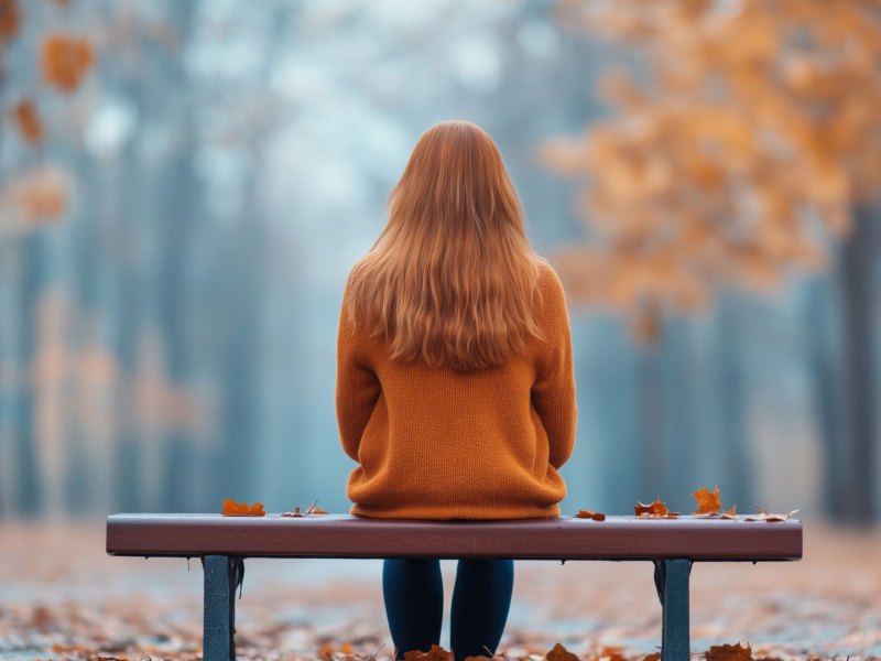 Frau mit roten Haaren und roter Jacke sitzt auf einer Bank in einem herbstlichen Wald