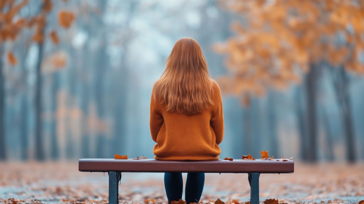 Frau mit roten Haaren und roter Jacke sitzt auf einer Bank in einem herbstlichen Wald